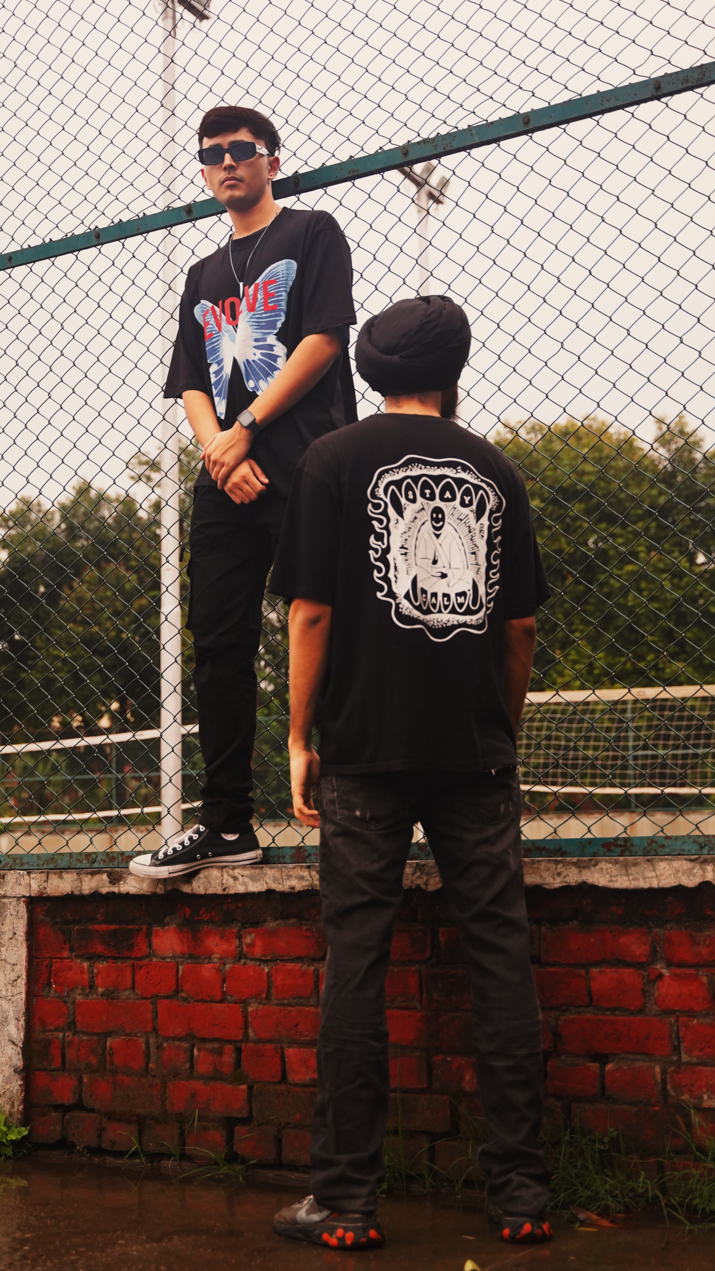 Two young men showcase stylish graphic T-shirts in a casual urban setting.
